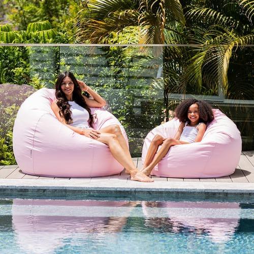Two women sitting on pink bean bags by a pool with greenery in the background